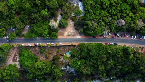 vista aérea de drones a vista de pájaro de una pequeña carretera rodeada de árboles tópicos y autos estacionados con la playa de praia do madeiro cerca de pipa con turistas, sombrillas y arena dorada en el norte de brasil