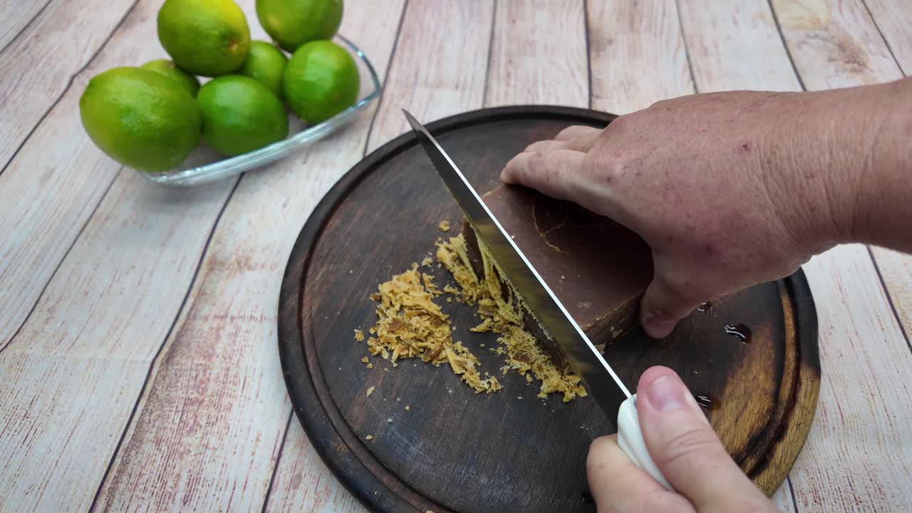 클로즈업된 손이 부엌칼을 사용하여 어두운 나무 판자 위에서 파넬라(정제되지 않은 사탕수수 블록)를 자르고 깎고 있어요. 전통 음료 준비를 위해 신선한 녹색 라임이 배경에 보이는군요.