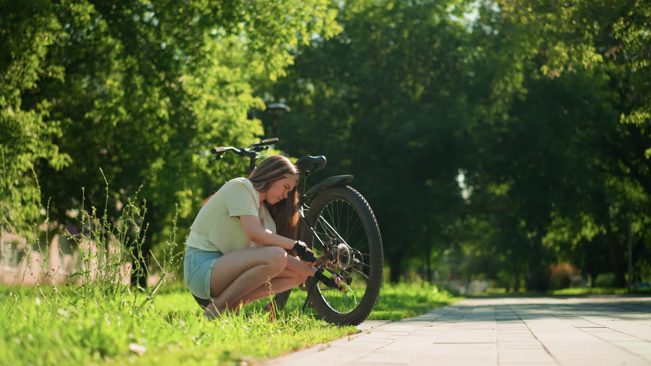 단호한 여성 사이클리스트는 빛이 비치는 잔디길에 누워서 활기찬 초록색과 나무로 둘러싸인 자전거의 뒷타이어에 공기를 프합니다.