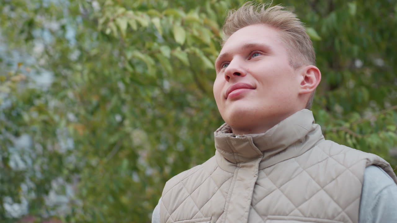 White man kneels with gentle smile on face during heartfelt moment surrounded by soft green foliage and swaying leaves in wind wearing quilted jacket