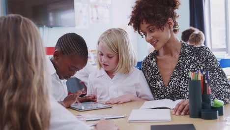 Female-Teacher-With-Two-Elementary-School-Pupils-Wearing-Uniform-Using-Digital-Tablet-At-Desk