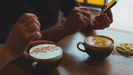 couple sitting at table in coffee shop looking at mobile phone together with foam art drinks