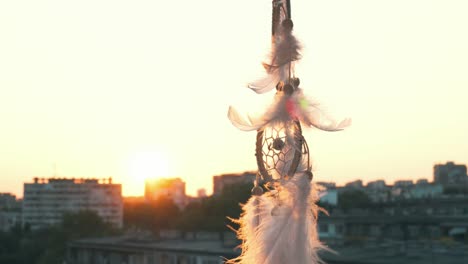 amulet dreams catcher waving in a light wind against the backdrop of a bright sunset and a bokeh of city lights. white light feathers rise in the wind. the disc of the setting sun. ethnic amulet