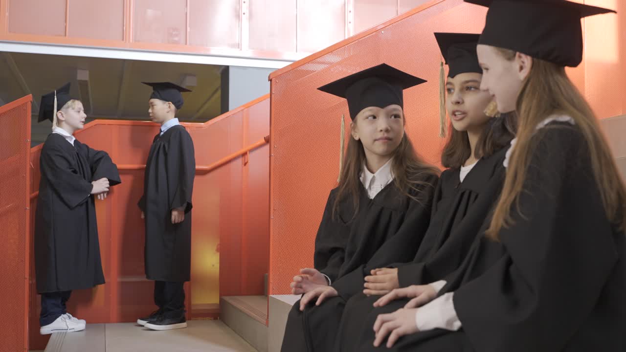 유치원 졸업식에서 모자와 가운을 입은 세 명의 행복한 어린 소녀들이 계단에 앉아 급우들에게 인사를 합니다