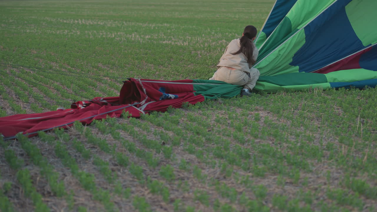 woman kneeling on vast green farmland pulling deflated multicolour hot air balloon canopy toward waiting wicker basket under gentle dusk sky