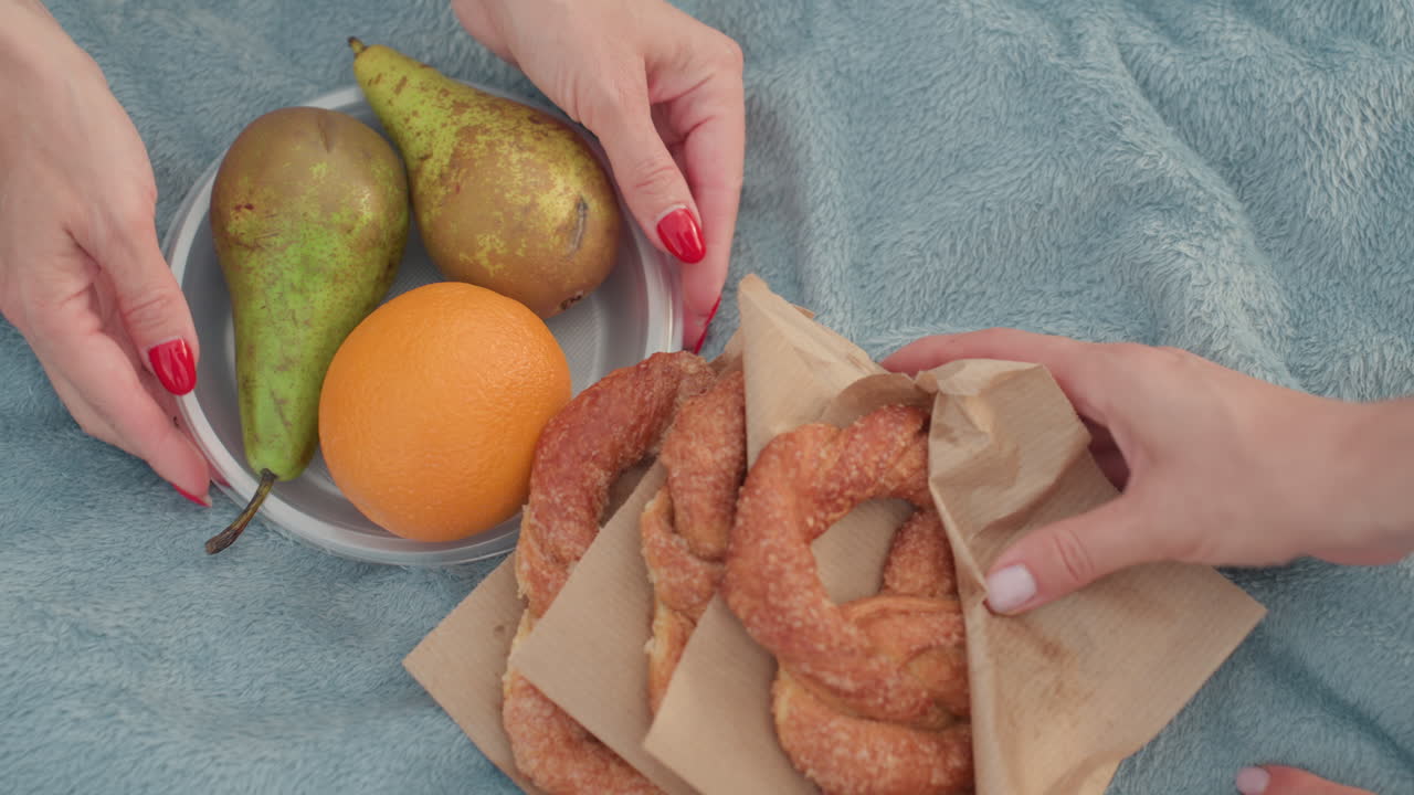 high angle view of female hands placing baked bread and plate of orange on picnic mat, soft blanket on grass, summer picnic preparation, warm light, casual sharing, healthy snack setup
