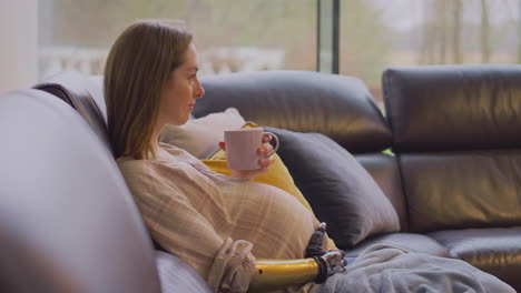 pregnant woman with prosthetic arm relaxing sitting on sofa at home with hot drink