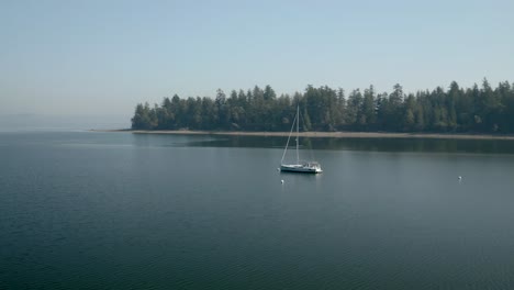 Aguas-Serenas-De-Mayo-Cove-Con-Un-Velero-Cerca-Del-Frondoso-Bosque-En-El-Parque-Estatal-Penrose-Point-En-Lakebay,-Estado-De-Washington,-Estados-Unidos