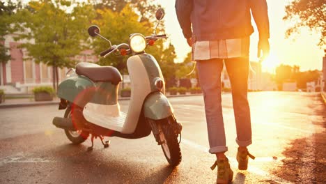 Close-up-shot-of-a-guy-in-denim-walking-up-to-his-green-moped,-getting-on-it-and-driving-off-on-the-street-during-sunrise-in-summer.-The-man-with-curly-hair-in-a-helmet-sits-on-a-moped-during-sunny-weather-in-summer