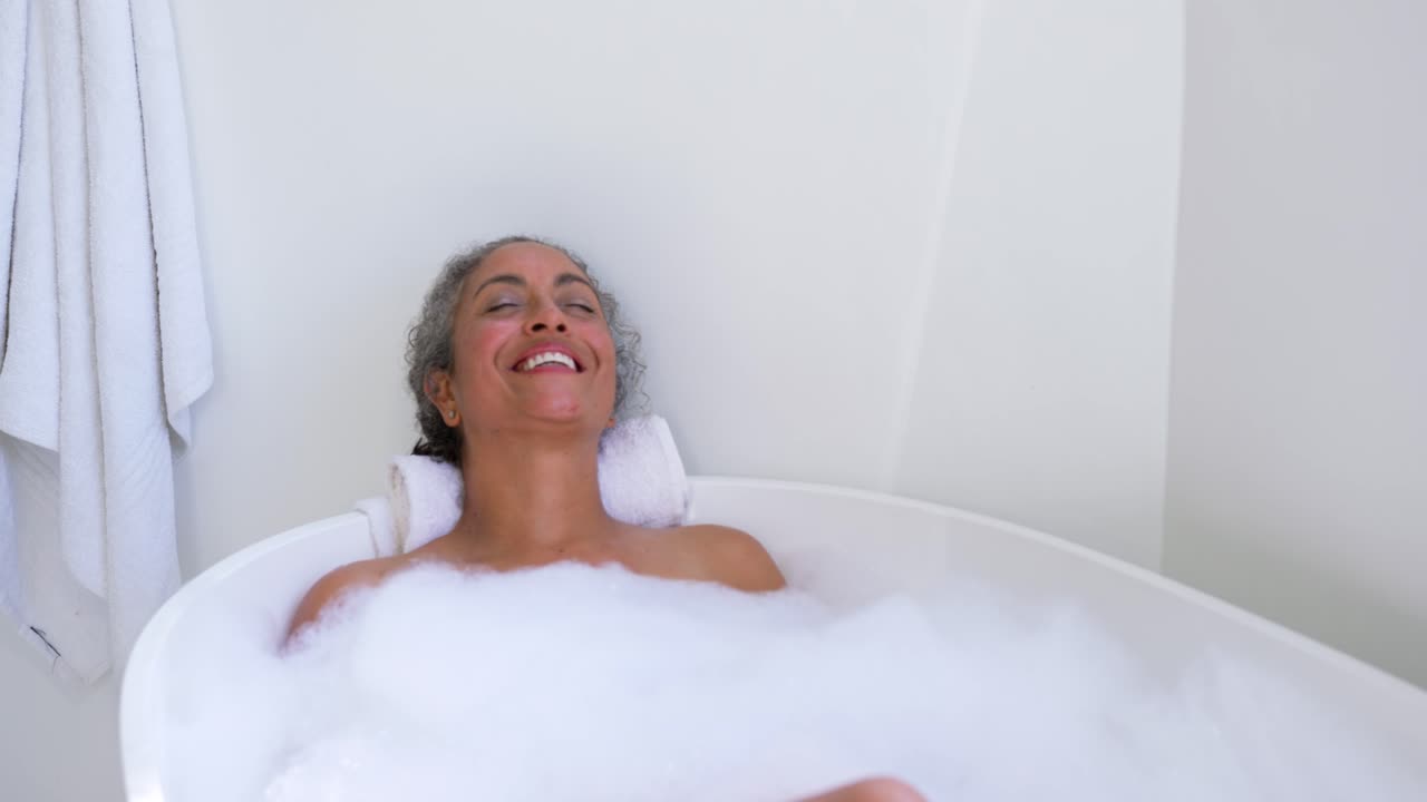 Senior African American woman relaxing in freestanding tub, smiling, breathing, knee nudging foam