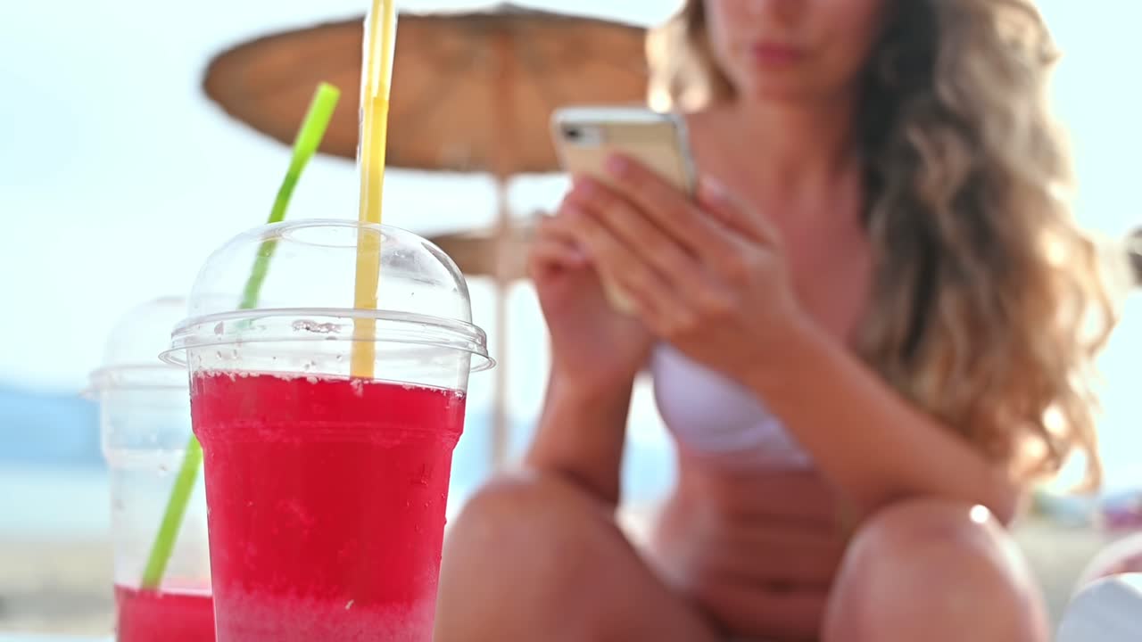 A red cocktail on the beach. Caucasian woman with smartphone sitting on the sunbed on the background. Sea, slow motion