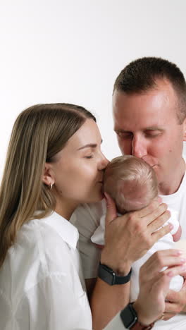 caucasian family of three. happy parents hold their little newborn and kiss him together. white backdrop. vertical video