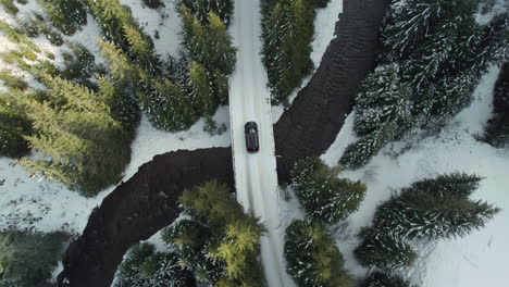 aerial view of a black car on a snowy road crossing over a bridge above a mountain river, surrounded by tall fir trees illuminated by sunlight