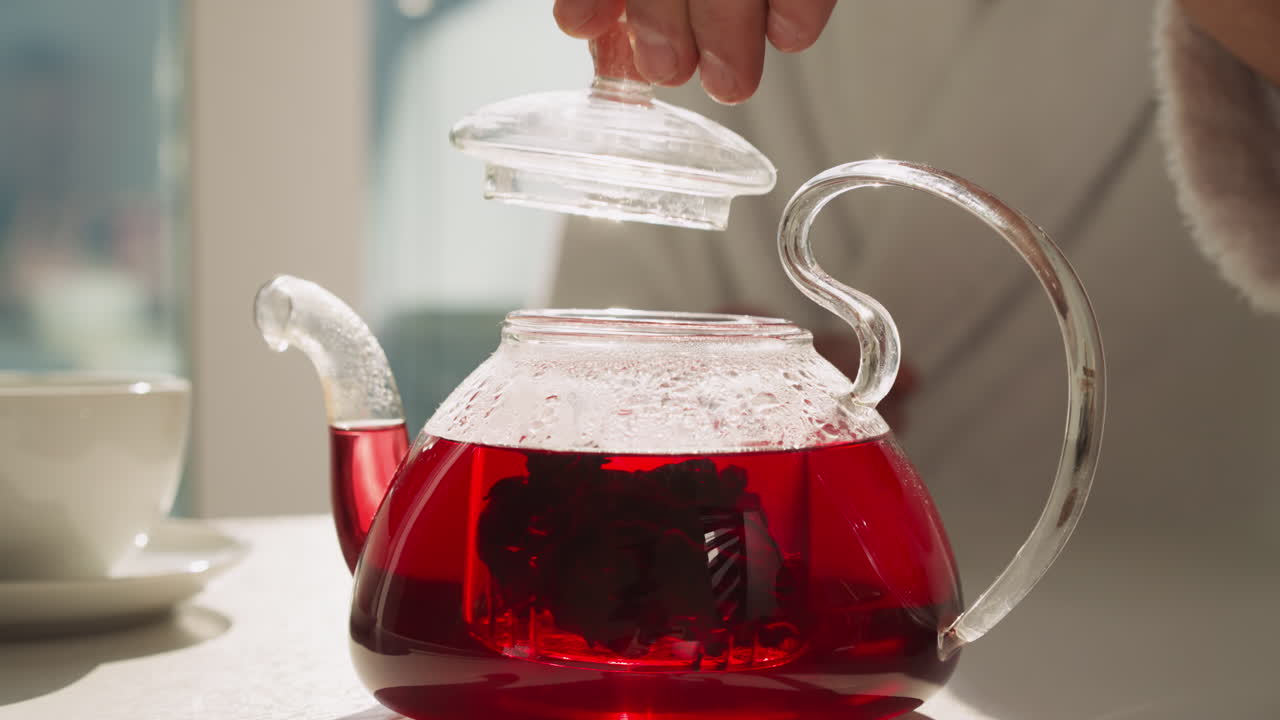 Man carefully puts cap on teapot with brewing tea in kitchen. Process of brewing hibiscus healthy tea according to traditions. Morning tea-time ritual