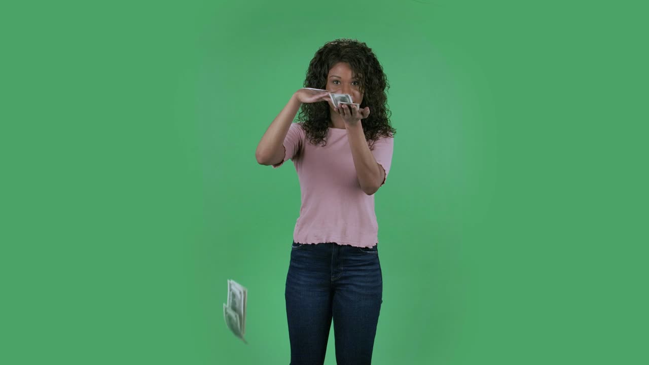 Portrait of beautiful african american young wealthy woman is scattering dollars with proud haughty expression, boasting rich life. Burning brunette with wavy hair in jeans and a beige blouse on a green screen in the studio