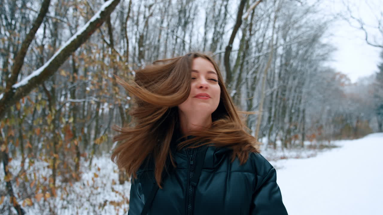Brunette woman wearing black blazer stands in winter park. Lady waves her long hair from side to side.