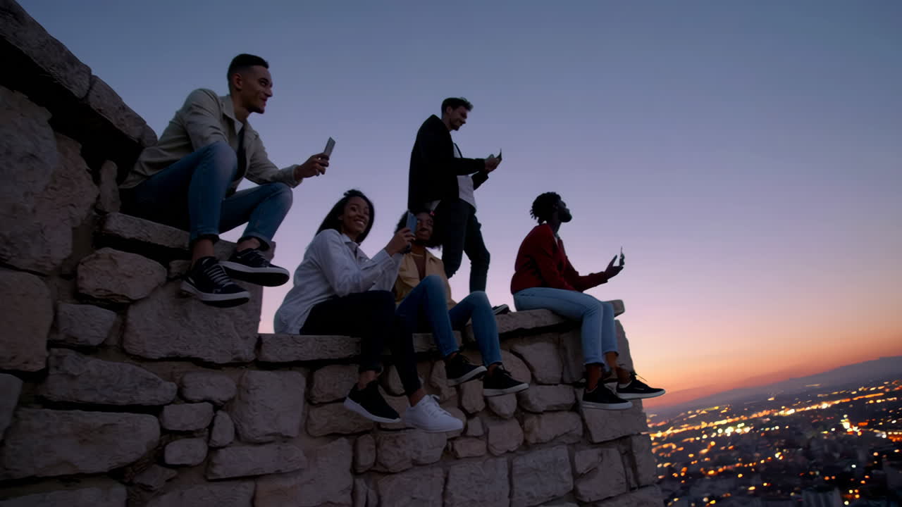 Diverse Group of Friends Engaged with Phones Overlooking City Lights at Dusk