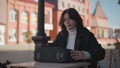 lady in black coat opens black bag while seated outdoors at wooden table in urban setting, background features blurred red brick buildings and a pole