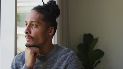 thoughtful biracial man with dreadlocks in hair bun looking out of window in living room