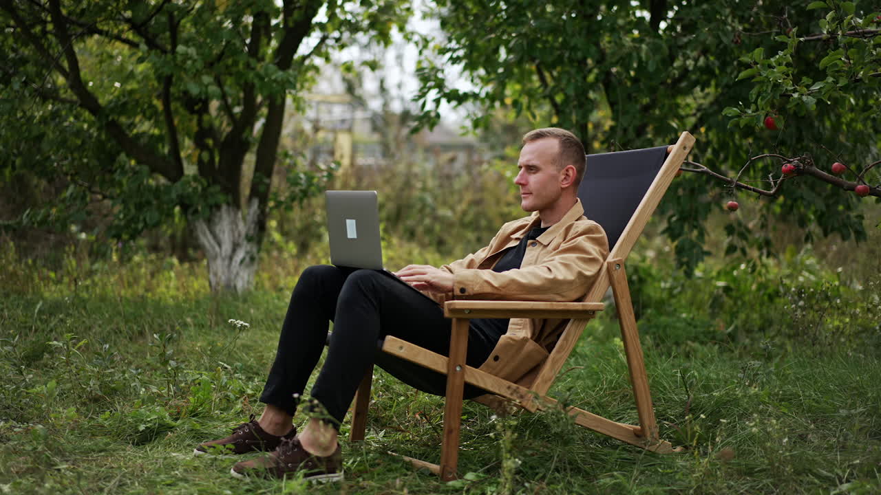 Busy man focused on his work on laptop sitting in garden. Camera slowly approaching freelancer who finishes work, closes laptop and leans back in his chair.