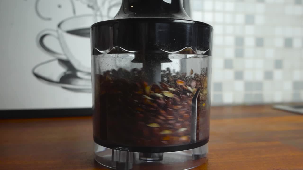 Brown seeds in a electrical blender spinning and being mixed in slow motion on a brown kitchen countertop table. The blender is in focus and background has coffee cup art and tiles slightly blurred.