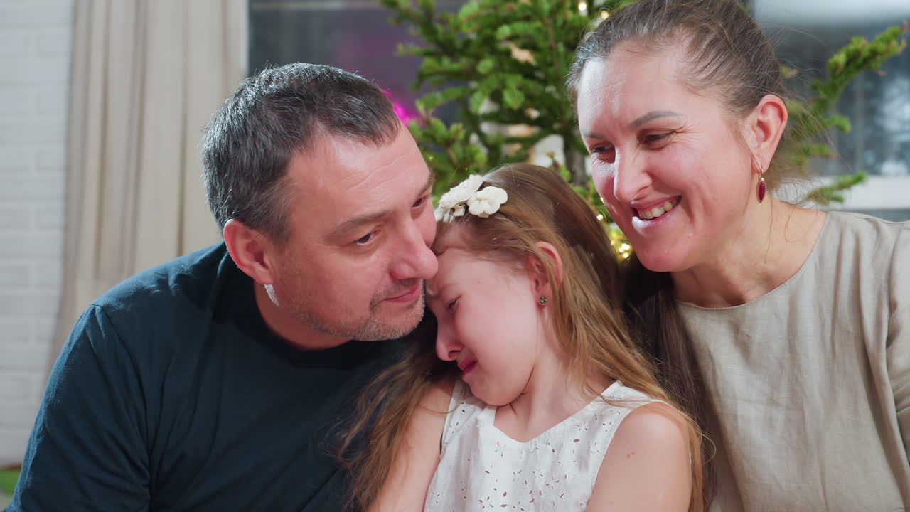 Close up view of loving dad trying to kiss young crying daughter on cheek while smiling mom looks on warmly during touching family moment near christmas tree inside cozy festive decorated home