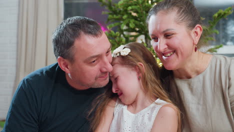 close up view of loving dad trying to kiss young crying daughter on cheek while smiling mom looks on warmly during touching family moment near christmas tree inside cozy festive decorated home