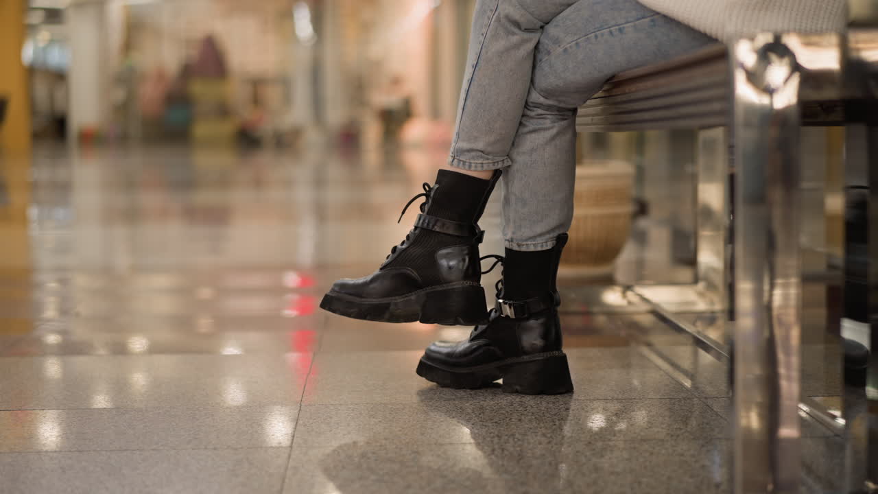 cross leg of stylish lady seated on mall bench wearing chunky black boots and denim jeans, showcasing casual fashion detail inside bright shopping mall under ceiling lights, captured from side angle