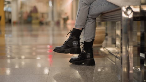 cross leg of stylish lady seated on mall bench wearing chunky black boots and denim jeans, showcasing casual fashion detail inside bright shopping mall under ceiling lights, captured from side angle