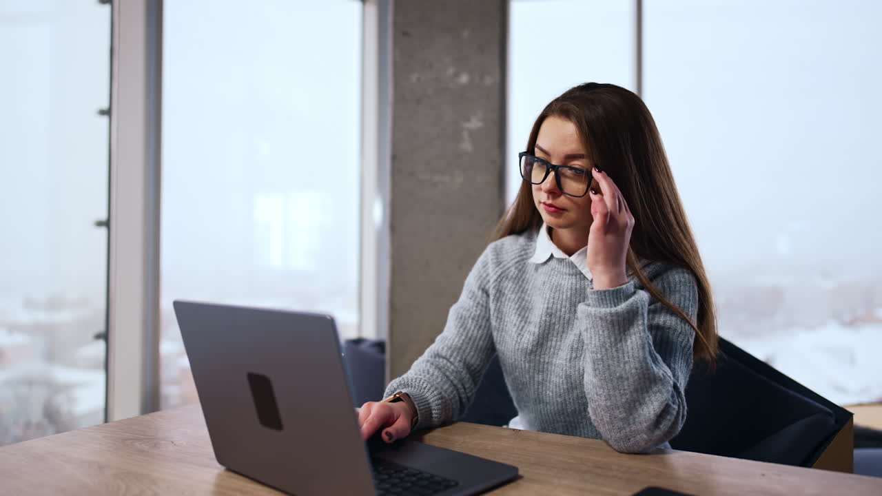 Woman working at laptop in the modern spacious office. Lady typing on keyboard stops suddenly and looks at the screen at surprise touching her eyeglasses.