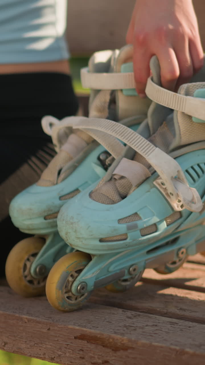 Close up of young lady dropping cyan roller skates on wooden bench as she sits beside it, abdomen slightly exposed while relaxed pose and blurred greenery background