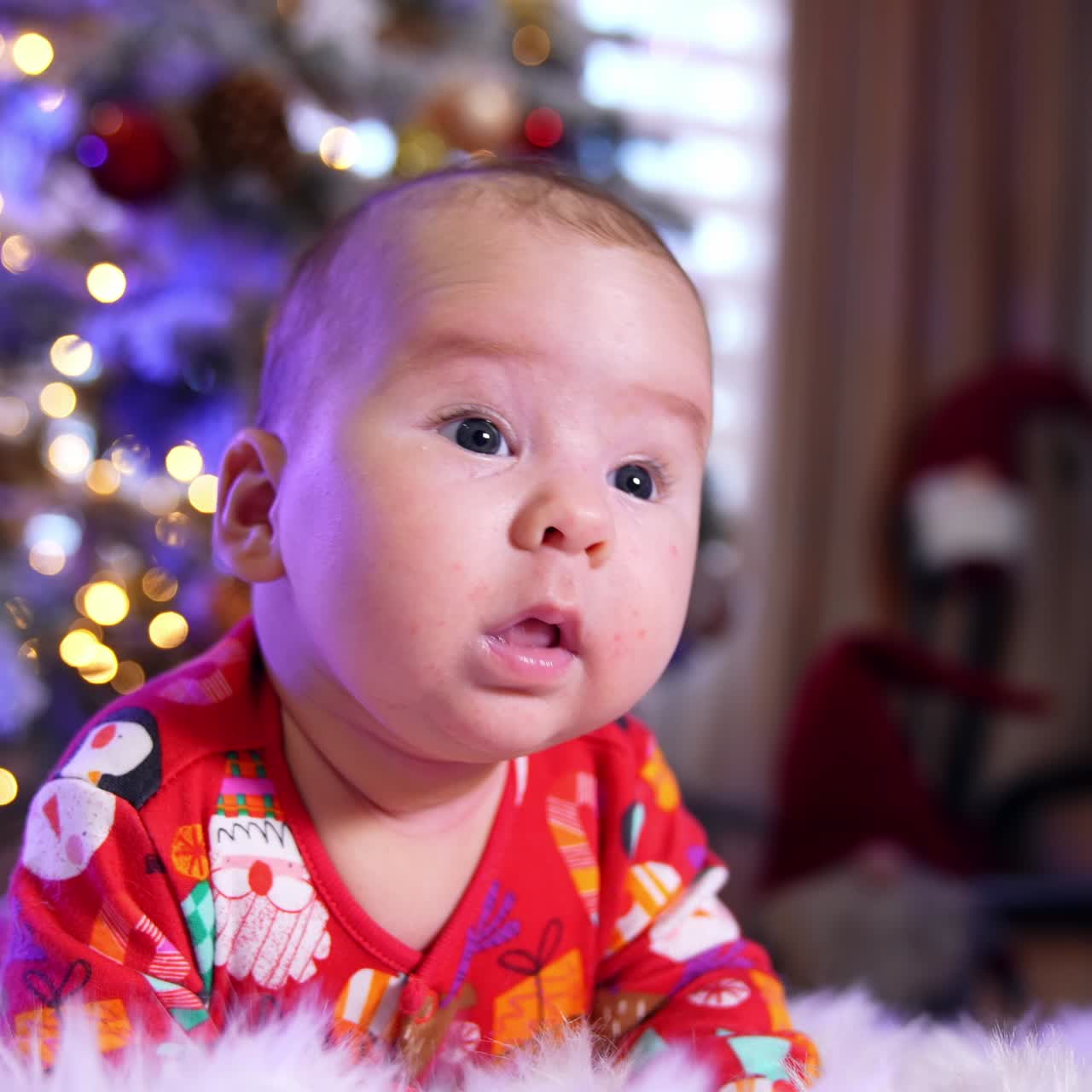 Beautiful child lies near Christmas tree on a plaid. Little infant kid looks up and then falls to his side raising his hands up