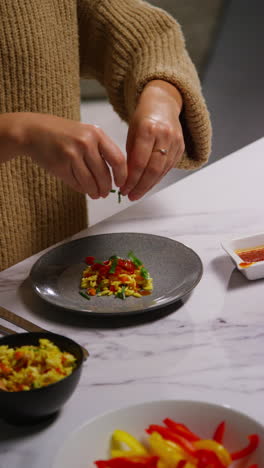 vertical video close up of woman at home in kitchen preparing healthy vegetarian or vegan meal sprinkling herbs onto orzo pasta and roasted tomatoes