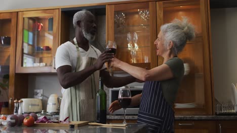 Caucasian-senior-woman-wearing-apron-giving-wine-glass-to-her-husband-in-the-kitchen-at-home