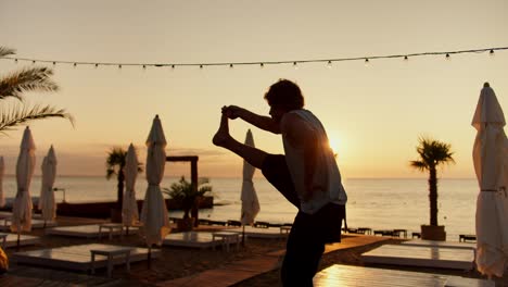 a young guy lifts his leg up and stretches on a sunny sale in the morning. doing sports and yoga on sunny beach in the morning. healthy lifestyle