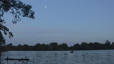 Toma-Panorámica-De-La-Luna-Al-Anochecer-Sobre-Un-Lago-Tranquilo-En-Un-Parque-Con-árboles.