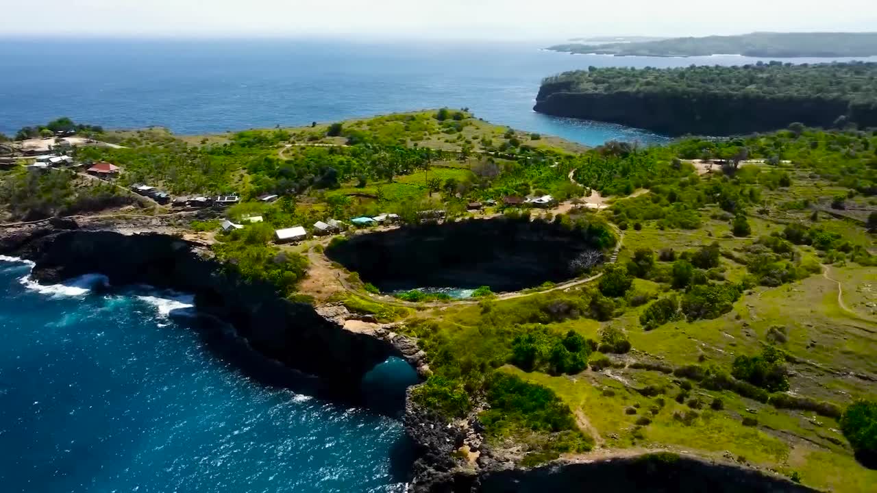 Aerial drone flying around in a circle or spinning around Bali Broken Beach land cavity and steep cliff shoreline during a sunny day. Landscape has small houses and ocean water waves moving below.