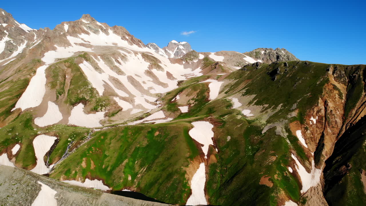 Valle alpina stagionale con chiazze di neve, crinali illuminati dal sole e prati verdi, ripresa da drone