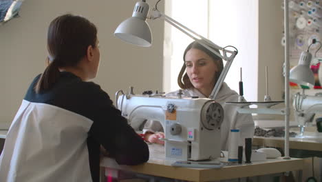 Professional-Caucasian-dressmaker-and-young-seamstress-choosing-fabric-and-talking-while-colleague-using-sewing-machine-in-designer-studio.