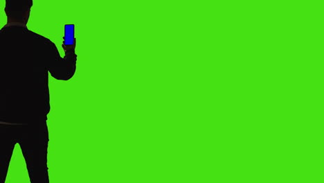 rear view of silhouetted man holding blue screen mobile phone towards camera standing against green screen background