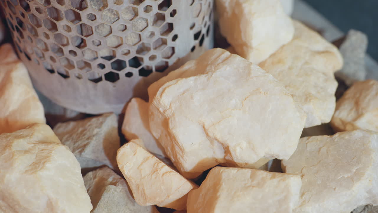 Top down close up of rough pale and dark sauna stones stacked around circular metal heater with hexagonal perforation pattern, highlighting natural textures