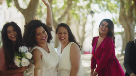 happy brides with guests lifting arms and looking at camera while celebrating wedding at the park