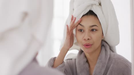 video of portrait of smiling biracial woman with towel on hair looking in mirror in bathroom