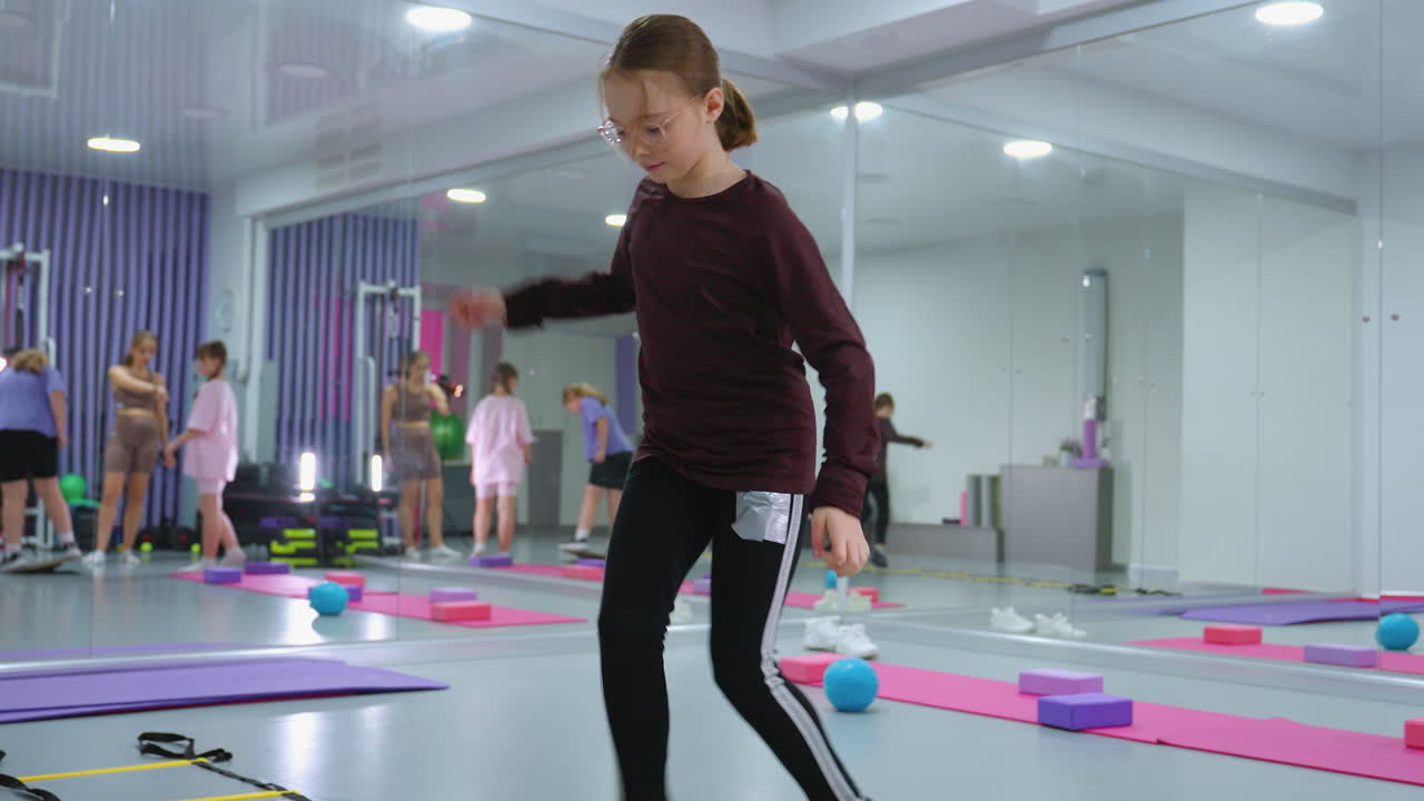 스피드 사다리에서 활력을 키우기 위해 민함을 연습하는 어린 소녀, 다른 사람들은 체육관에서 다양한 피트니스 활동을 하며, 운동 장비와 참가자를 반영하는 큰 거울을 가지고 있습니다.