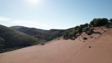 aerial dolly above sloping sand dune with grassy mountain side behind, footsteps across landscape