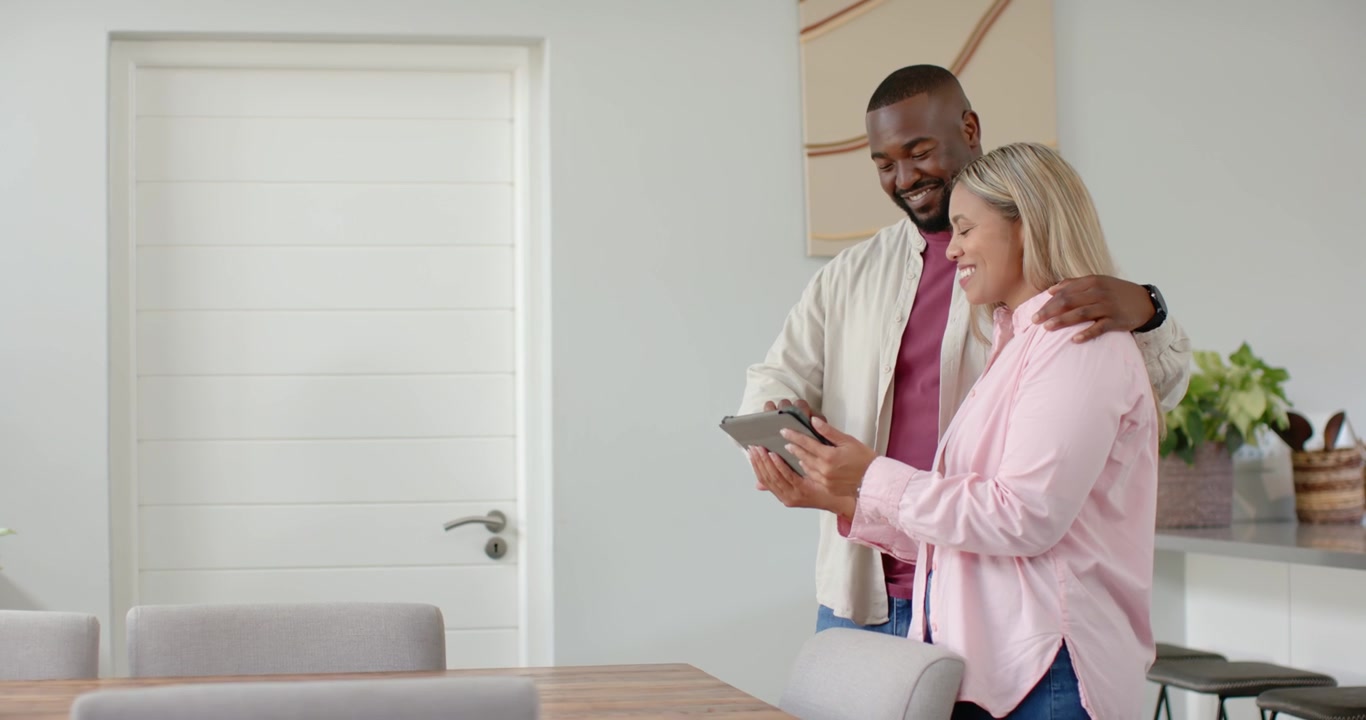 Coppia afroamericana che tiene un tablet e naviga tra i contenuti nella cucina di casa, appoggiata e sorridente.