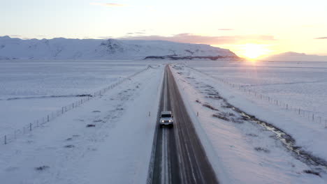 Antenne:-Nahaufnahme-Des-Jeeps,-Der-Auf-Islandstraße-Mit-Schneeweißen-Bergen-Und-Sonnenuntergangsschnee-Fährt,-Arktis