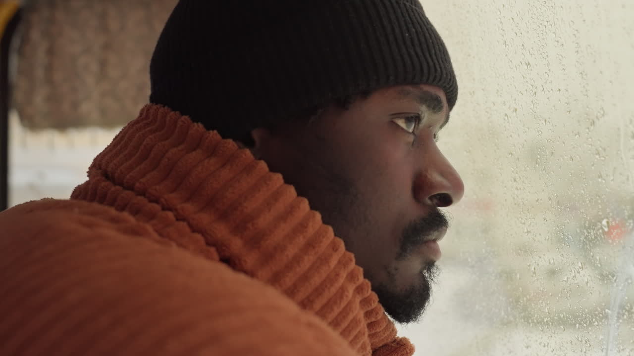 Side profile man gazing through window, closeup portrait of orange knit sweater and black beanie against rainy glass, focused expression conveying anticipation and quiet resilience during urban