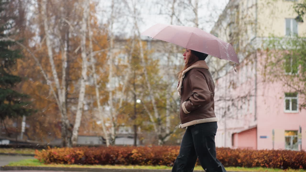 Cheerful girl happily running in light snowfall holding umbrella wearing knit cap brown shearling jacket black trousers one hand pocket surrounded by colorful autumn leaves on calm overcast day