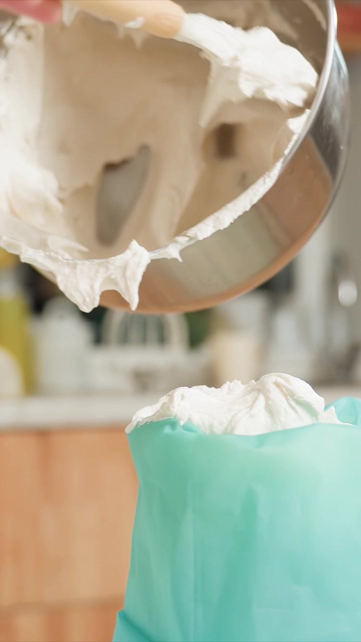 Primo piano di una persona con un top verde che versa glassa bianca densa. La sostanza cremosa riempie un sac à poche mentre una spatola la raschia via. Ambiente cucina con mobili in legno